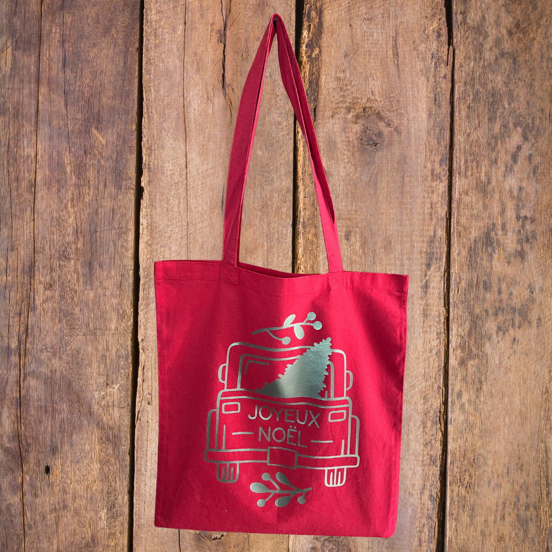 Tote bag rouge avec une image ecrit Joyeux Noël posé sur un fond en bois.